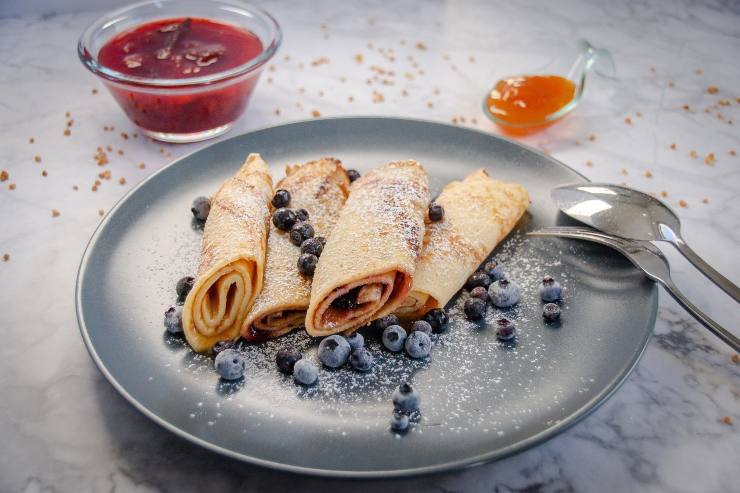 4 rotolini di crepes su un piatto con attorno dei mirtilli e dietro delle ciotoline con delle salse