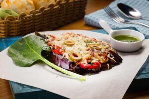fetta di melanzana sopra ad un piatto con sopra pomodoro, formaggio e olive e accanto una ciotolina con dentro una salsa