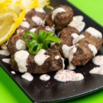 polpette su un piatto nero ricoperte da una salsina bianca con accanto una fettina di limone