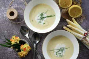 due piatti pieni di vellutata con accanto degli asparagi bianchi, dei fiori e dei limoni