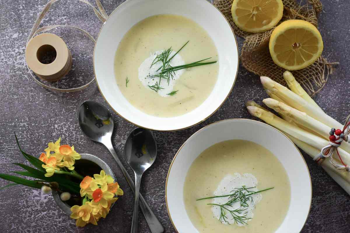 due piatti pieni di vellutata con accanto degli asparagi bianchi, dei fiori e dei limoni