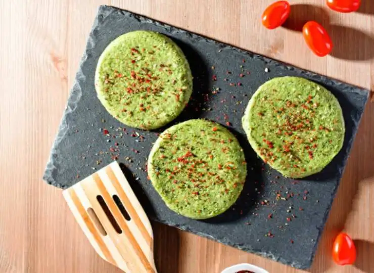 tre burger di broccoli sopra ad un tagliere nero e accanto un mestolo e dei pomodorini