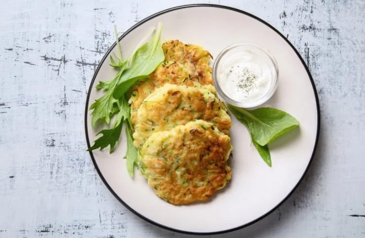 tre frittelle di zucchine su un piatto con accanto delle foglie di insalata e una salsina bianca