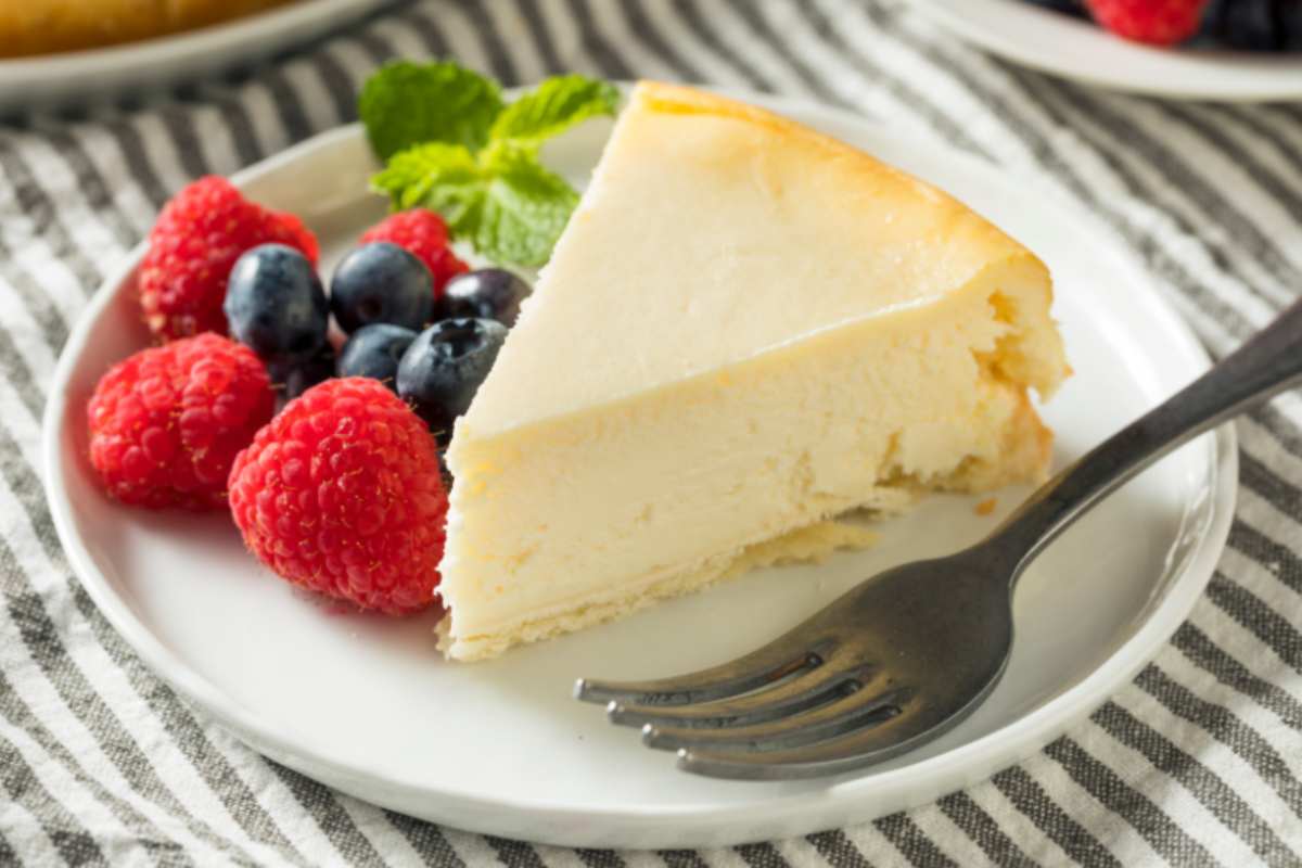 fetta di torta su un piatto con accanto fragole, mirtilli e foglie di menta e una forchetta
